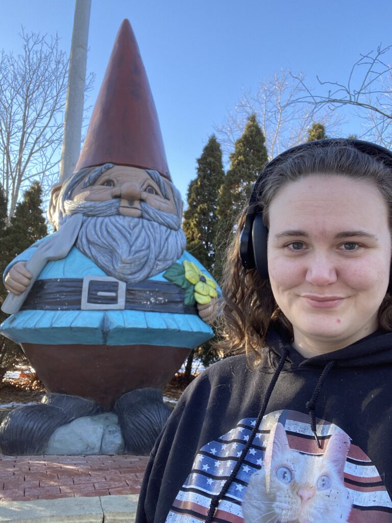 me, a white woman wearing headphones standing in front of the world's largest concrete gnome taking a selfie.